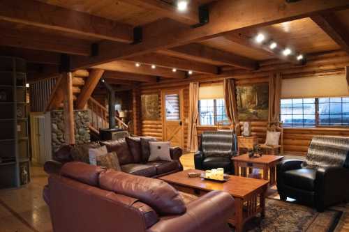 Cozy log cabin interior with wooden beams, leather sofas, a coffee table, and large windows letting in natural light.