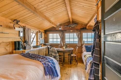 Cozy wooden cabin interior with two beds, a dining table, and large windows overlooking a scenic view.