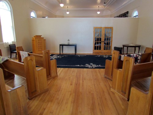 Interior of a simple, empty room with wooden pews, a table, and a rug, featuring large windows and warm lighting.