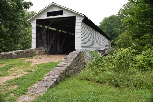 A white covered bridge with a stone foundation, surrounded by greenery and trees.