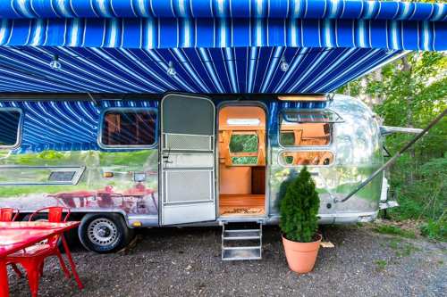 A shiny silver Airstream trailer with a blue striped awning, surrounded by greenery and red outdoor chairs.