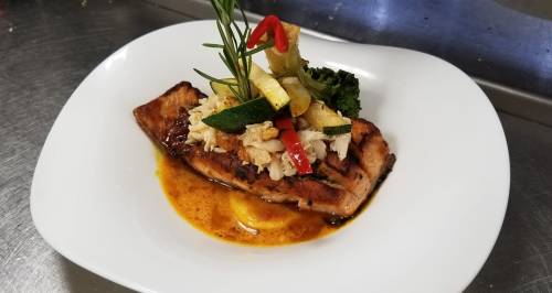 Grilled salmon fillet topped with crab meat, garnished with vegetables and herbs on a white plate.