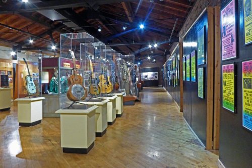 A music museum display featuring guitars in glass cases and colorful posters on the walls, with wooden flooring.