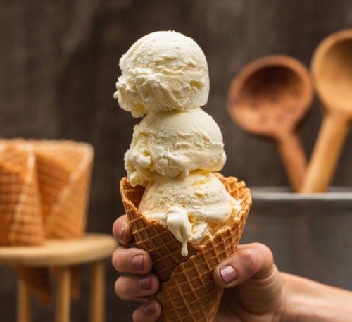 A hand holding a waffle cone with three scoops of vanilla ice cream, with wooden spoons in the background.