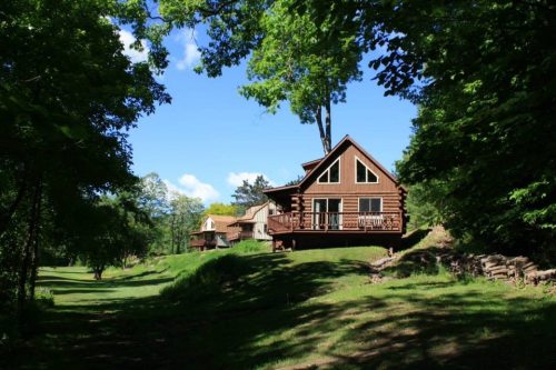 A log cabin surrounded by trees, with a clear blue sky and green grass in the foreground.