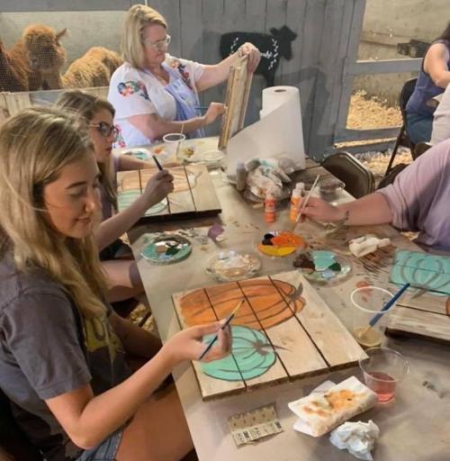 A group of people painting on wooden boards at a table, with art supplies and a farm setting in the background.