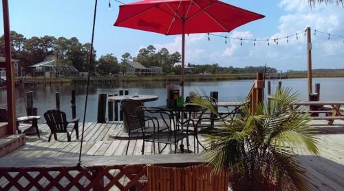 A scenic dock with a red umbrella, chairs, and a view of calm water and trees under a blue sky.