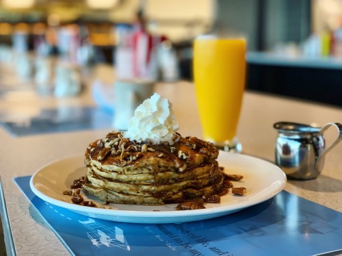 A stack of pancakes topped with whipped cream and nuts, served with a glass of orange juice and syrup on the side.