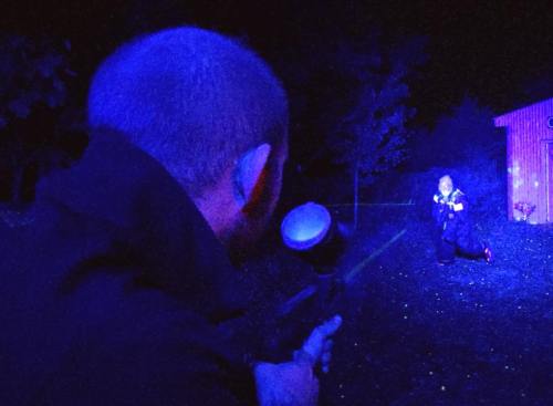 A person aims a paintball gun at another person crouching in a dark, blue-lit outdoor setting.