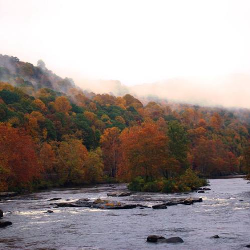 A serene river flows through a vibrant autumn landscape with colorful trees and misty mountains in the background.
