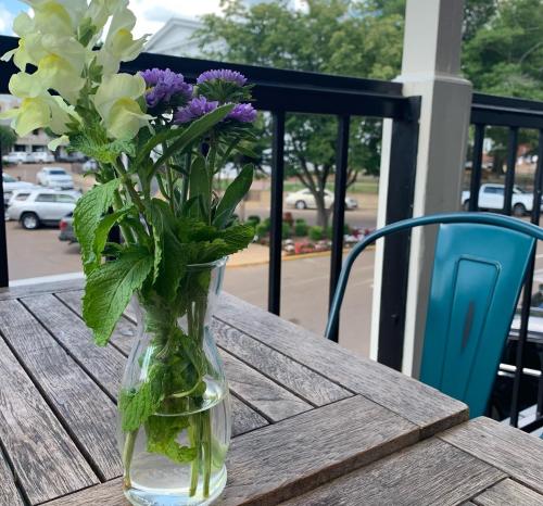 A small vase with mixed flowers sits on a wooden table, with a view of a street and parked cars in the background.