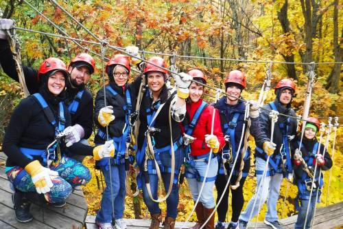 A group of people in helmets and harnesses smiles while zip-lining through a colorful autumn forest.