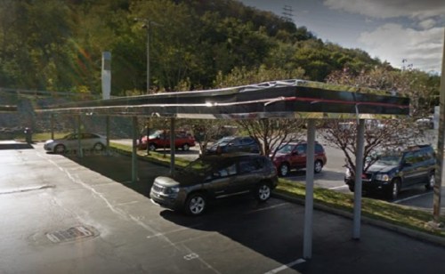 A parking lot with several cars under a metal canopy, surrounded by trees and a hillside in the background.