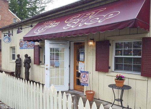 A quaint gift shop with a maroon awning, open sign, and potted plant, surrounded by a white picket fence.