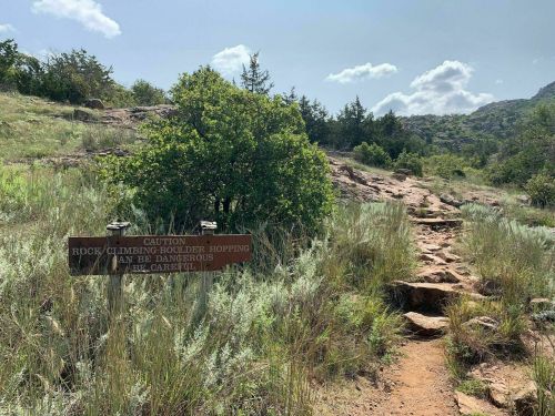 A rocky trail leads through grassy terrain, with a caution sign about boulder climbing and hopping ahead.