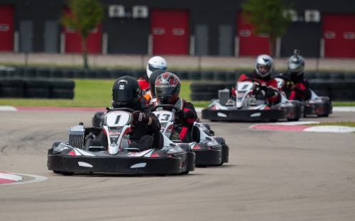 A group of go-karts racing on a track, with drivers wearing helmets and racing suits.