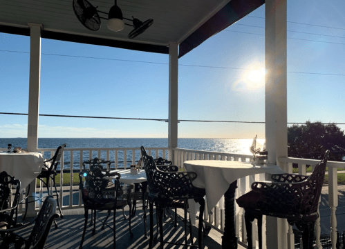 A seaside restaurant patio with tables overlooking the ocean and a bright sun reflecting on the water.