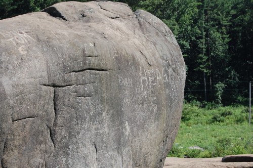 A large rock surface covered in various carvings and graffiti, surrounded by greenery and trees in the background.
