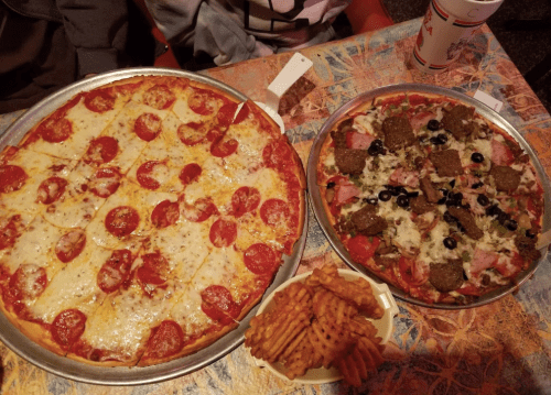 A table with two pizzas: one pepperoni and one with various toppings, alongside a serving of waffle fries.
