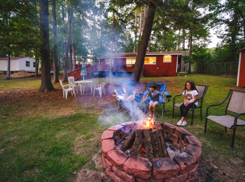 A cozy campfire scene with three people sitting around a fire pit in a wooded area, surrounded by chairs and cabins.