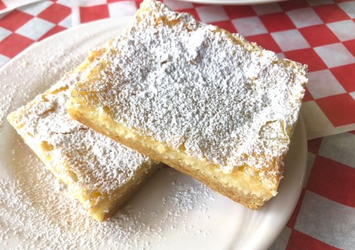 Two powdered sugar-dusted dessert bars on a white plate, resting on a red and white checkered tablecloth.