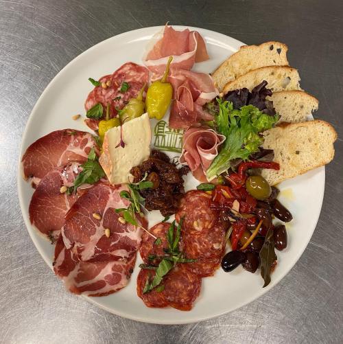 A platter featuring assorted cured meats, cheeses, olives, pickles, and fresh greens, served with slices of bread.
