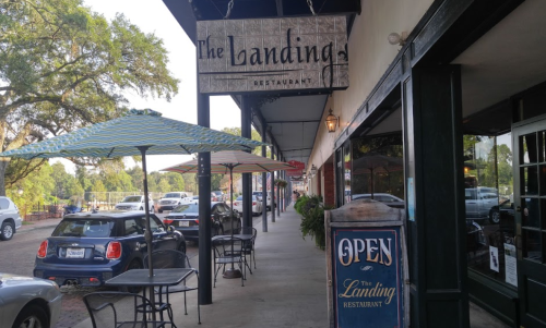 A view of The Landing restaurant with outdoor seating, umbrellas, and an "Open" sign on a sunny day.