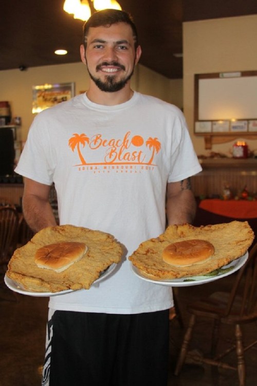A man in a "Beach Blast" shirt holds two large plates of food, featuring oversized fried chicken and a burger.