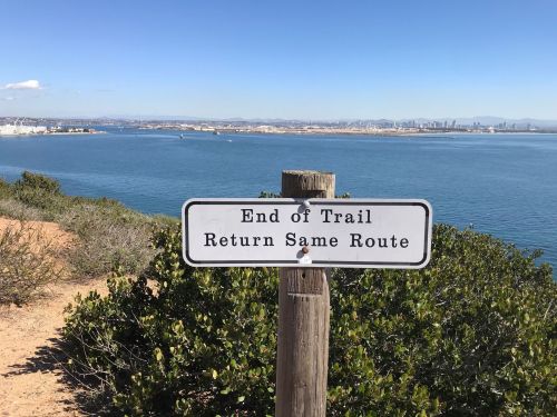 Signpost at a scenic viewpoint, indicating "End of Trail" with instructions to "Return Same Route" beside a calm bay.
