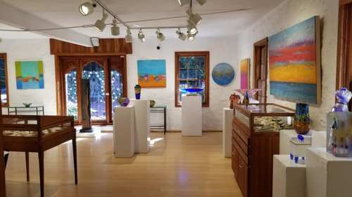 Art gallery interior featuring colorful paintings and glass sculptures displayed on white pedestals and wooden furniture.
