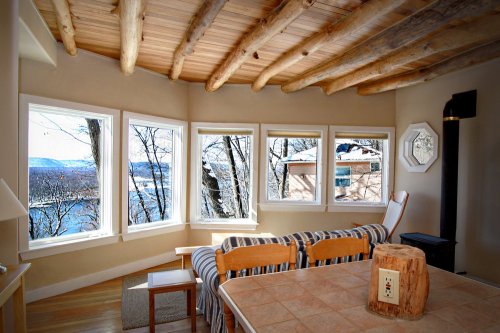 Cozy interior with wooden beams, a dining table, and large windows showcasing a scenic view of trees and a river.