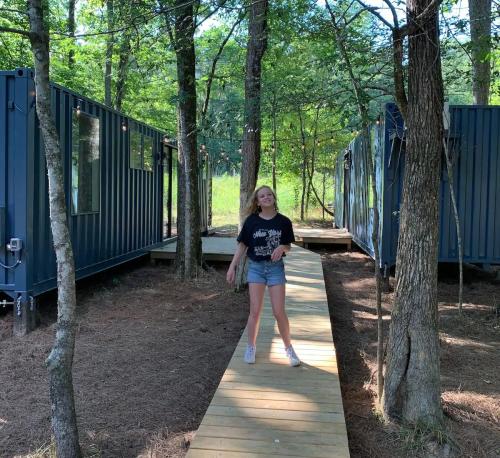 A girl stands on a wooden path between two blue shipping container cabins in a wooded area.