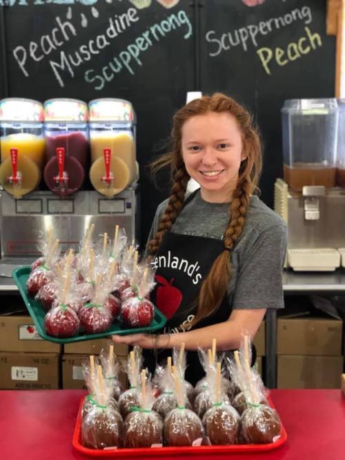 A smiling young woman with braids holds a tray of caramel apples in a candy shop, with drink dispensers in the background.