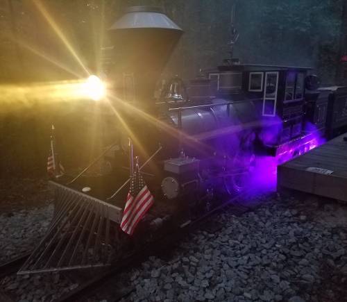 A vintage steam locomotive with glowing purple lights and American flags, surrounded by mist in a wooded area.