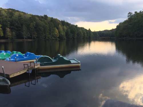A serene lake at sunset with colorful paddle boats docked and surrounded by lush green trees.