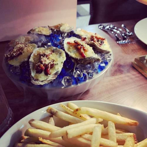 A plate of baked oysters topped with breadcrumbs and bacon, served on ice, alongside a bowl of French fries.