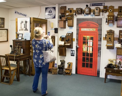 A woman takes a photo in a museum displaying vintage telephones, including a red phone booth.