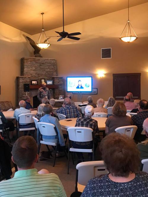 A speaker presents to an audience in a spacious room with a stone fireplace and a large screen displaying a video.