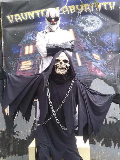 A creepy clown stands behind a skeleton in a dark cloak, set against a haunted house backdrop with a full moon.