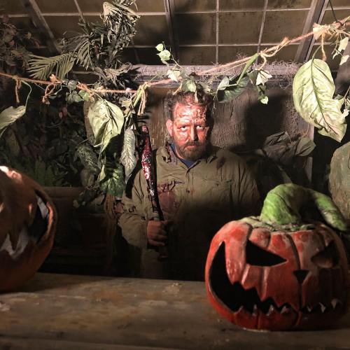 A man with a bloody face holds a weapon, surrounded by eerie foliage and carved pumpkins in a dimly lit setting.