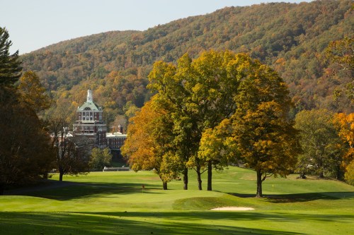A scenic view of a golf course with vibrant autumn trees and a historic building in the background, surrounded by mountains.