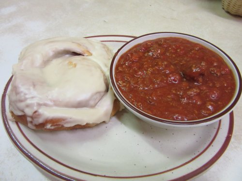 A plate with a large cinnamon roll and a bowl of chili, both served on a white background.