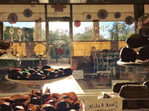 A view through a shop window showcasing a display of assorted chocolates and pastries, with sunlight streaming in.