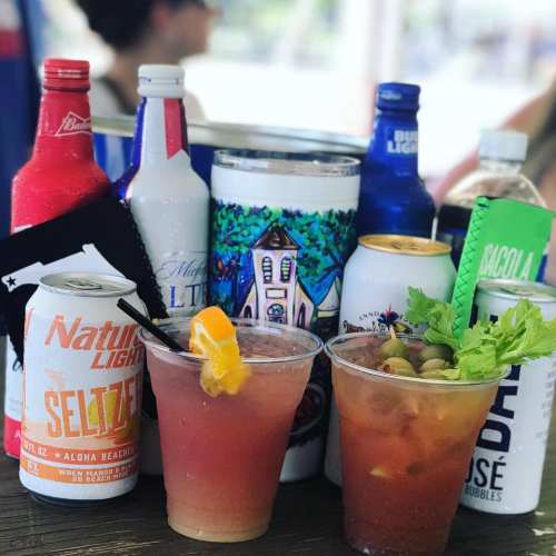 Two cocktails with garnishes sit in front of various colorful beverage cans and bottles on a wooden table.