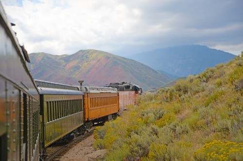 A vintage train travels along a scenic route with mountains and colorful foliage in the background.