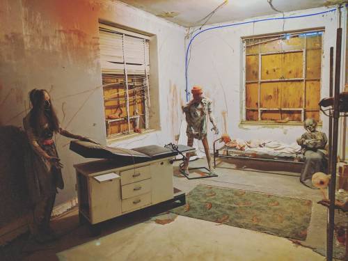 A dimly lit, eerie room with creepy mannequins, a medical table, and stained walls, creating a horror atmosphere.