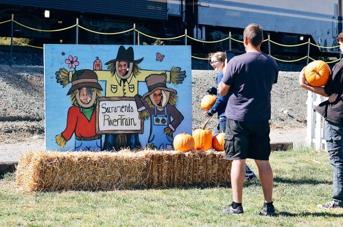 A colorful mural of scarecrows with people posing behind it, surrounded by pumpkins and hay bales at an outdoor event.
