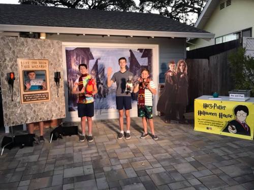 Three kids in summer clothes pose in front of a Harry Potter-themed Halloween house backdrop with props.