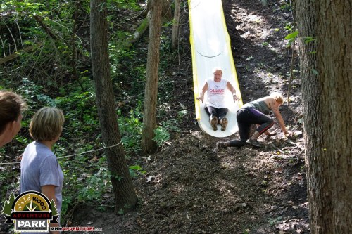 Two people on a slide in a wooded area, one sliding down while the other is climbing up from the ground.