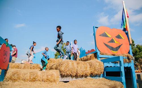 Children playing on a haystack structure at a festive outdoor event, with a large painted pumpkin in the background.